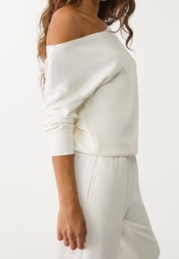Sweat-shirt blanc à épaules dénudées en tissu doux, avec manches longues et poignets côtelés, assorti à un pantalon de jogging assorti.