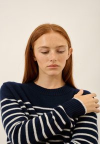 Granatowy sweter z dzianiny z białymi paskami, z prążkowanym kołnierzem i mankietami. Materiał wydaje się miękki, z dbałością o detale w szyciu i luźnym kroju.