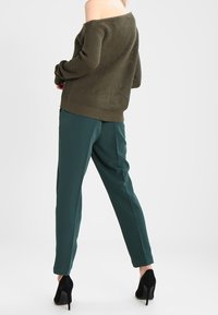 Oliwkowy sweter z dekoltem off-shoulder w prążki, zestawiony z zielonymi spodniami typu tapered i czarnymi butami na obcasie, prezentujący luźny krój i kontrast tekstur.