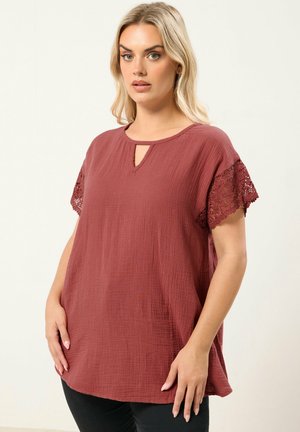 Blonde Frau, die ein lockeres, strukturiertes weinrotes Oberteil mit Schlüsselloch-Ausschnitt und Spitzenärmeln trägt, vor einem einfarbigen hellen Hintergrund stehend.