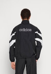 Svart vindjacka med vita accentlinjer, elastiska ärm- och halsavslut. Har "adidas"-logga i vitt på baksidan. Lättviktig tyg.