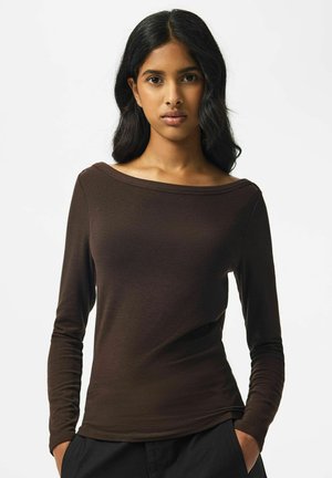 Femme aux longs cheveux foncés portant un haut marron à manches longues et un pantalon noir, les mains dans les poches, debout devant un fond blanc.