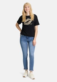 Zwarte T-shirt met een grafisch logo van lippen in luipaardprint en tekst. Gecombineerd met blauwe skinny jeans en witte sneakers. Korte mouwen, relaxed fit.