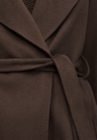 Gros plan d’un manteau en laine marron avec un large revers et une ceinture en tissu nouée à la taille.