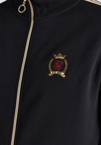 Chaqueta negra con cremallera y alto cuello, con un logotipo y una corona bordados en oro y rojo, tela suave y detalles de cremallera en contraste.