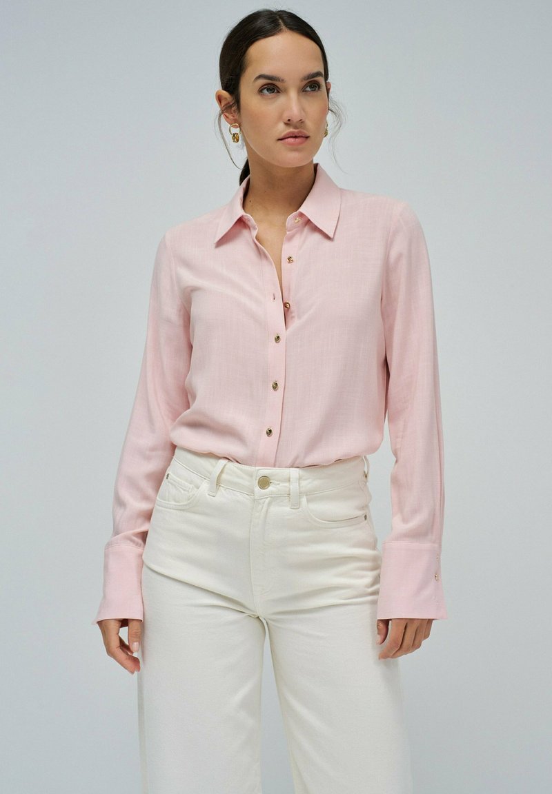 Camisa de manga larga rosa claro con frente abotonado y botones en tono dorado, combinada con jeans blancos de cintura alta. Textura de tela suave.