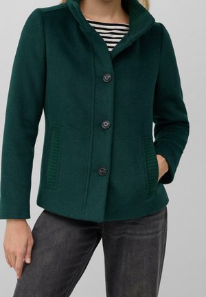 Femme portant une veste en laine verte foncée boutonnée avec des poches côtelées et une chemise rayée noire et blanche en dessous, associée à un jean gris foncé.