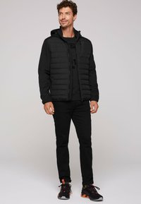 Mustat pehmustetut takki, jossa on huppu, vetoketjukiinnitys ja sivutaskut, päällä musta graafinen t-paita, mustat farkut ja urheilukengät.