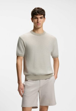 Junger Mann, der einen beige farbenen, kurzärmligen Strickpullover und hellgraue Shorts trägt, steht mit einer Hand in der Tasche vor einem weißen Hintergrund.