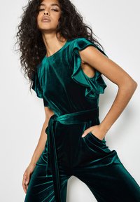 Mono de terciopelo verde esmeralda con mangas con volantes, cinturón anudado y bolsillos laterales; textura suave con un ligero brillo.