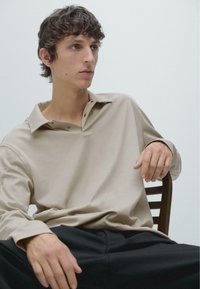 Beige poloshirt met lange mouwen en een zachte textuur, met een kraag en twee knopen, gecombineerd met een donkere broek, zittend op een houten stoel.