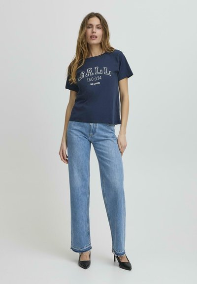 Tricou din bumbac bleumarin cu inscripția albă "BALL 1974 THE JEANS" pe față, asortat cu blugi largi de culoare albastru deschis și tocuri negre.