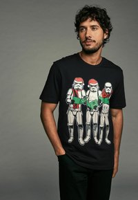 Černé tričko s motivem tří stormtrooperů v Santa kloboucích, v bílé a červené barvě, kteří drží zelený objekt, navržené pro každodenní nošení.