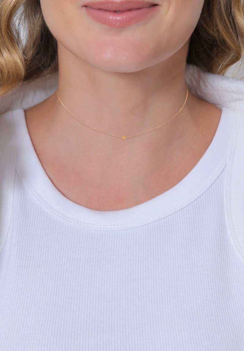 Goldfarbener Choker mit einer zarten, dünnen Kette und einem kleinen Sternanhänger, getragen mit einem weißen gerippten Oberteil.