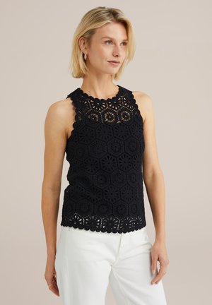 Schwarzes Häkel-Top mit hexagonalem Muster, gewelltem Ausschnitt und strukturiertem Finish, kombiniert mit weißen Hosen.
