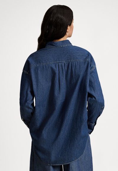 Sieviete ar gariem tumšiem matiem valkā brīvi krītošu tumšu džinsa krekls un bikses, stāvot ar muguru pret kameru pret vienkāršu fonu.