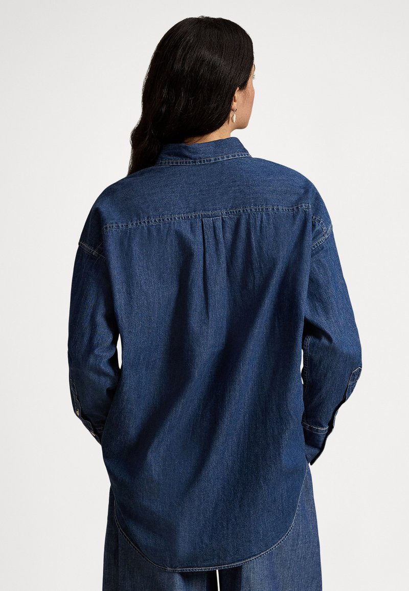 Donna con capelli lunghi e scuri indossa una camicia e pantaloni ampi di denim scuro, in piedi con la schiena rivolta verso la fotocamera su uno sfondo semplice.