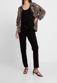 Top negro sin mangas combinado con pantalones negros holgados y una chaqueta abierta estampada que presenta tonos rosa, dorado y negro. Sandalias negras de tiras completan el conjunto.