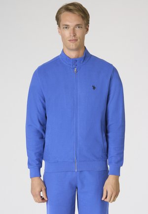 Giovane uomo che indossa una giacca blu con zip e colletto alto e pantaloni blu abbinati, in piedi davanti a uno sfondo bianco semplice.