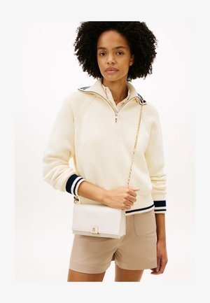 Žena s kučeravými vlasmi, oblečená v krémovom svetri s námorníckymi pruhmi, béžovými krátkymi nohavicami a drží bielu kabelku cez rameno so zlatým reťazovým remienkom.