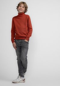 Maglione a collo alto rosso, jeans slim-fit grigi e sneakers bianche con accenti rossi. Tessuto a maglia texturizzato e vestibilità casual.