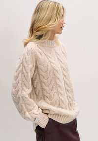 Gruby kremowy sweter w sploty warkoczowe z wysokim kołnierzem, opuszczonymi ramionami i prążkowanym dołem; noszony na ciemnej bordowej spódnicy.