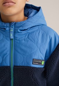 Kék steppelt kapucnis dzseki, tengerészkék polárbéléssel, kontrasztos zöld cipzárral és az elején téglalap alakú márkajelzéssel.