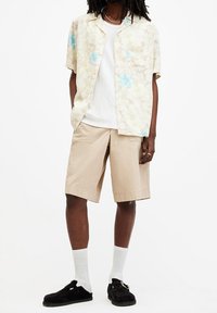 Uomo con una camicia corta beige a fiori sopra una canottiera bianca, pantaloni corti beige, calze bianche e mocassini neri, in piedi con le mani in tasca.