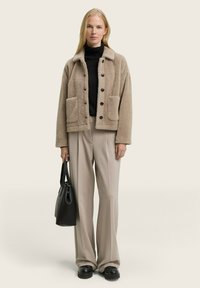 Beige fleece jack met een kraag, drie knopen aan de voorkant en twee opgestikte zakken, gecombineerd met lichte broek en een zwarte handtas.
