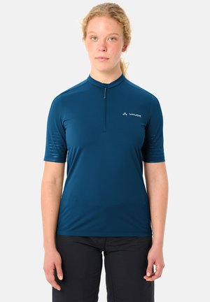 Femme aux cheveux bouclés clairs portant un t-shirt de sport turquoise à manches courtes avec une fermeture éclair et un pantalon noir, debout devant un fond blanc uni.