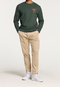 Grön crewneck sweatshirt med chiné-textur, beige tapered byxor och vita sneakers. Har en geometrisk grafik med sol och vågor.