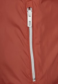 Tissu de veste couleur rouille avec une fermeture éclair argentée, d'une texture lisse et d'un petit tirette de marque.