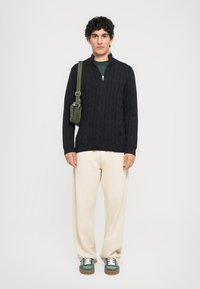 Άνδρας που φοράει μαύρο πουλόβερ με κοτσίδες, κρεμ παντελόνι, πράσινα αθλητικά παπούτσια και πράσινη τσάντα χιαστί, στέκεται μπροστά σε λευκό φόντο.