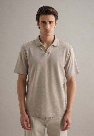 Uomo che indossa una polo beige a maniche corte e pantaloni chiari, in piedi contro uno sfondo neutro e semplice.