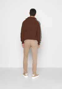 Man som står vänd bort med en brun tröja med gröna ränder på ärmarna, beige byxor och beige sneakers mot en enkel bakgrund.