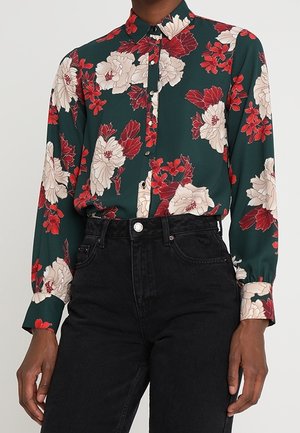 Zelená halenka s dlouhým rukávem a velkým červeno-bílým květinovým vzorem, zastrčená do vysokým pasem černých džínů.