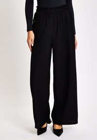 Black wide-leg pants with a gathered elastic waistband, two side pockets, and smooth fabric texture, paired with black pointed-toe heels.
