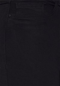 Närbild av svart denimtyg som visar den övre delen av jeansfickan med synlig sömnad och en liten svart nitar.