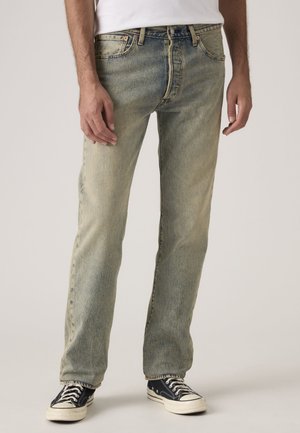 Mann trägt ausgewaschene blaue Straight-Leg-Jeans und schwarze Sneakers und steht vor einem einfarbigen Hintergrund.