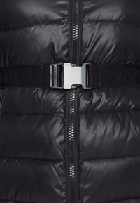 Svart vadderad väst med en glansig, slät yta, framdragkedja och ett svart justerbart bälte med ett metallspänne.