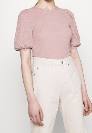 Vrouw draagt een getextureerde pastelroze top met pofmouwen, ingestopt in hoog getailleerde off-white broek met voorzakken en knoopsluiting.
