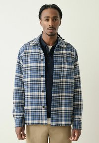 Blue and white plaid jacket with a collar, button front, and textured fabric, layered over a navy shirt. Beige pants complete the outfit.