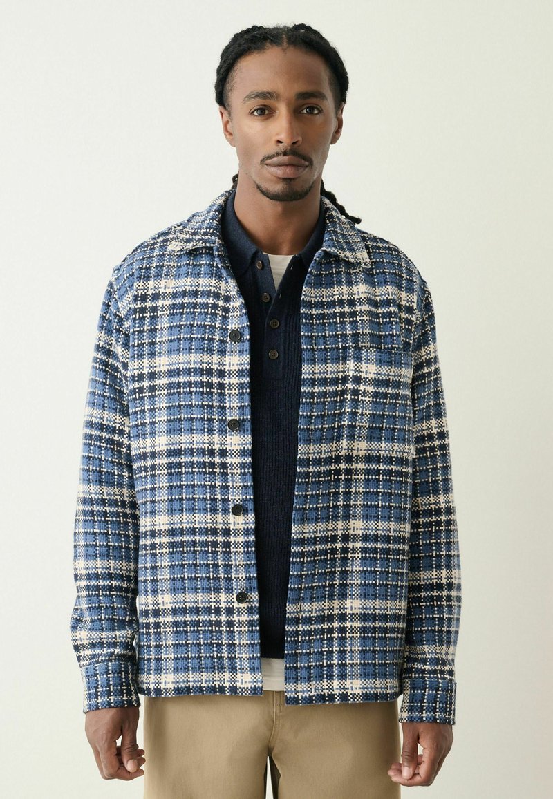Blue and white plaid jacket with a collar, button front, and textured fabric, layered over a navy shirt. Beige pants complete the outfit.