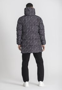 Chaqueta negra acolchada con un patrón de logotipos blancos repetidos, cuello alto con capucha y un diseño de longitud media. Se lleva con pantalones negros y zapatos blancos.