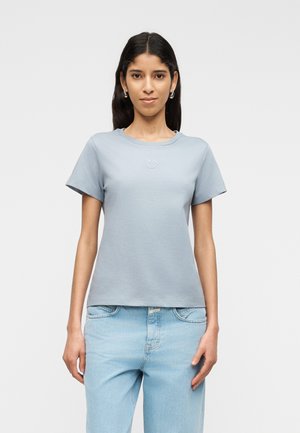 Fiatal nő hosszú sötét hajjal, világoskék rövid ujjú pólót és világoskék farmert visel, egy egyszínű háttér előtt állva.