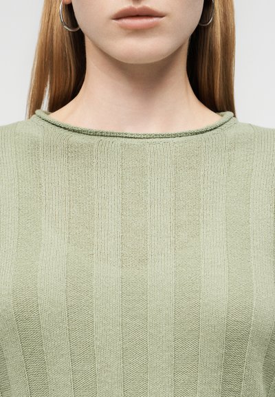 Γυναίκα που φοράει ασημένια σκουλαρίκια κρίκους και ένα ανοιχτό πράσινο πλεκτό πουλόβερ με ραβδώσεις και ελαφρώς γυρισμένο λαιμό.