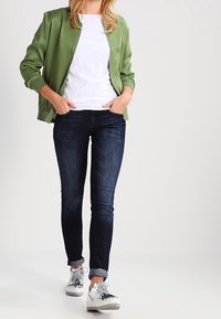 Veste aviateur verte portée sur un t-shirt blanc, associée à un jean skinny bleu foncé et des baskets argentées. La veste possède des poignets côtelés et une fermeture éclair à l'avant.