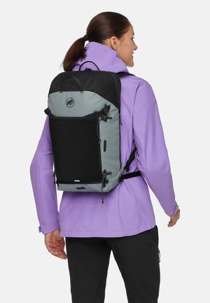 Kvinde iført lilla jakke og sort-grå rygsæk, der kigger til siden, stående mod en ensartet lys baggrund.