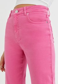 Jean en denim rose vif avec une taille haute, cinq poches et une braguette zippée. Présente une texture subtile et des détails métalliques brillants à l'avant.