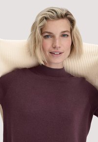 Bordeauxfarbener Pullover mit hohem Kragen; glatte Textur. Cremefarbener, gerippter Stoff teilweise im Hintergrund sichtbar. Model mit kurzen, hellen Haaren.